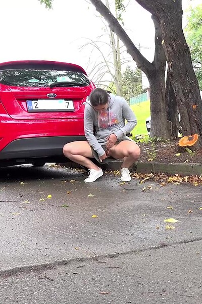 A desperate brunette squats behind a car and sprays a messy stream of piss onto the road.
